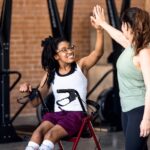 Woman sitting in wheelchair high fives female friend after exercising